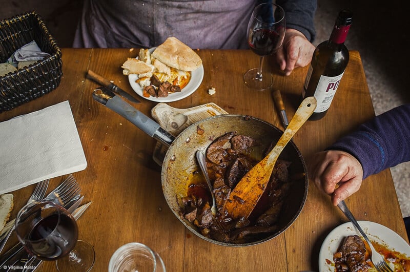 An overhead view of a wooden table featuring a cooked meat dish in a pan with a wooden spoon, surrounded by plates with bread and food remnants. Two people are holding glasses of red wine. A bottle of wine is on the table, along with a basket of bread and some utensils.