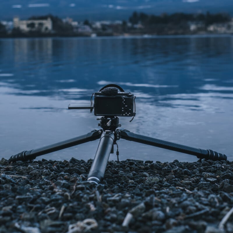 A camera mounted on a low tripod is positioned on a rocky shore facing a calm body of water. The distant shoreline and mountains are blurred in the background, creating a serene, dusk-like atmosphere.