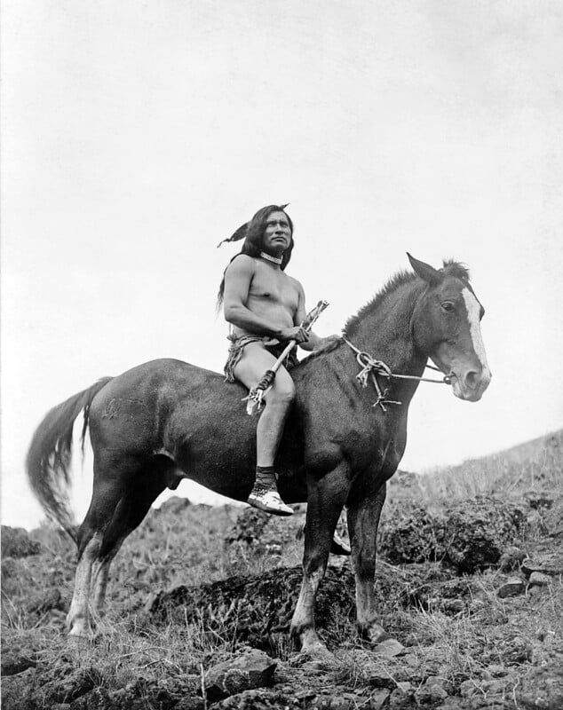 A black and white photo of a man with long hair, wearing minimal clothing and traditional footwear, riding a horse on rocky terrain. He holds a small staff or tool, and the horse has a white marking on its face. The background is a clear sky.