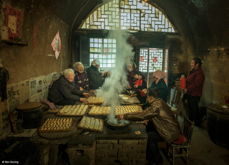 A group of elderly women are gathered in a traditional kitchen with rustic, earthen walls and decorative windows. They are making and steaming dumplings around a large table, with steam rising from a central cooking pot. The scene is cozy and communal.
