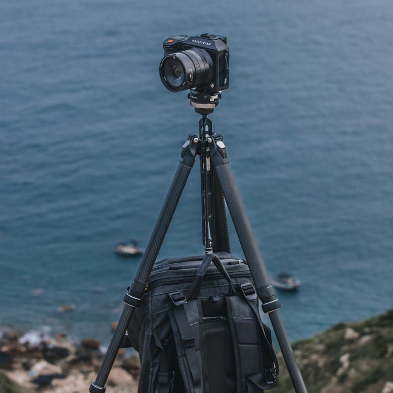 A camera mounted on a tripod stands on a cliff overlooking an expansive ocean. A black backpack is resting at the base of the tripod. The scene is calm with a slight blur of distant water and rocks visible below.