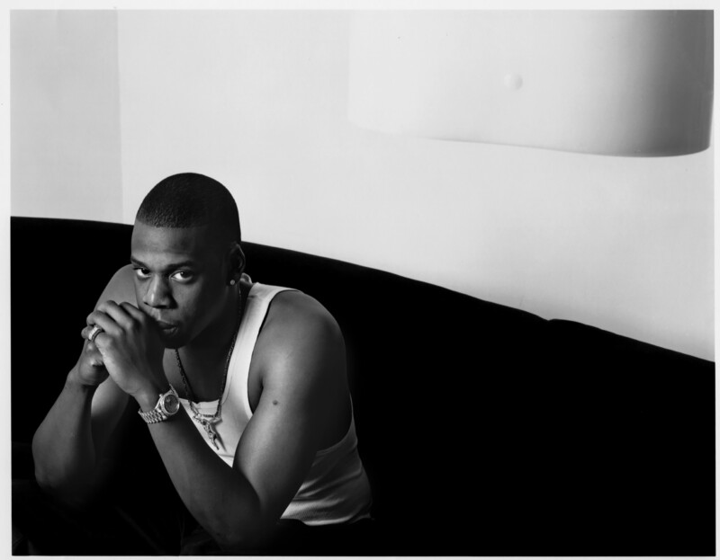 A person, seated on a dark couch, gazes intently at the camera. They wear a white tank top and a watch, with hands clasped near their mouth. The background is minimalistic, featuring a white wall with a large light fixture hanging from above.