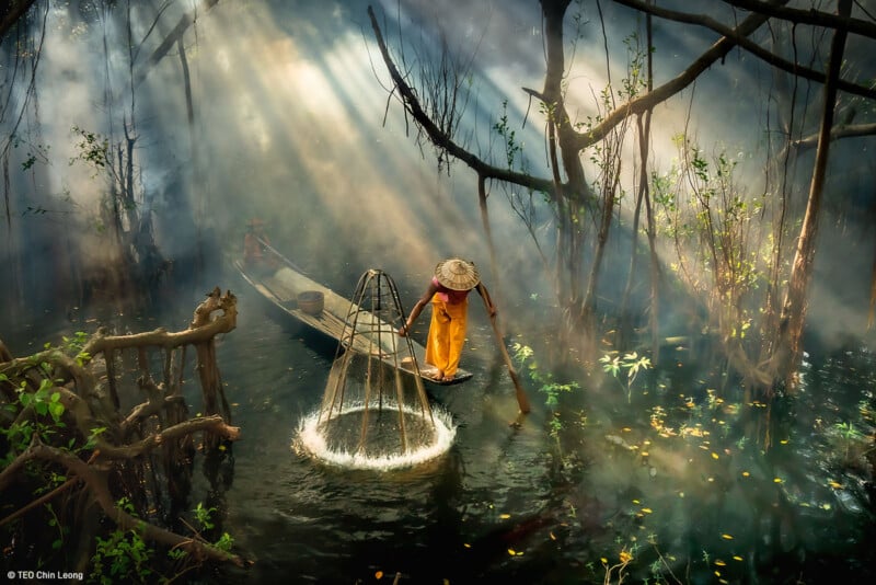 A person wearing a straw hat and bright orange clothing stands on a small wooden boat, using a net to fish in a misty, tree-covered waterway. Sunlight filters through the trees, creating an ethereal, serene atmosphere.