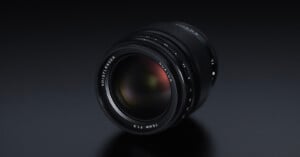 A close-up view of a Voigtländer Nokton 75mm f/1.5 lens resting on a dark surface. The lens features a sleek black design with white text around the front element indicating the brand, model, and aperture specifications.