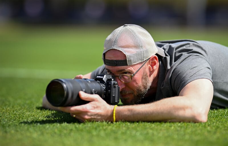 A man lies on the grass, wearing a backward cap and glasses, focusing on taking a photo with a Nikon camera. His elbows are propped on the ground, and he wears a grey shirt and a yellow bracelet. The background is blurred, highlighting his activity.
