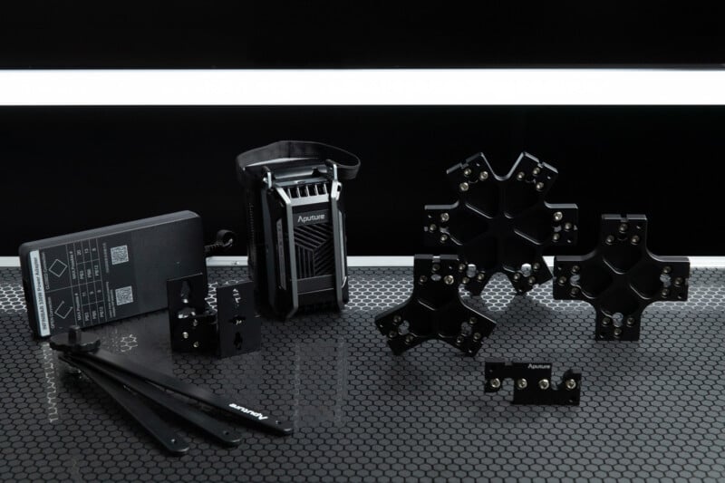 A collection of black Aputure lighting accessories is displayed on a hexagonal patterned table. The items include a control box, mounting plates, two straps, and various brackets, all arranged in an orderly manner with a black and white background.