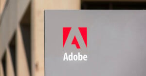 A close-up photo of an Adobe sign featuring the company's name and logo. The logo consists of a stylized red 'A' above the word 'Adobe' in white text, all set against a grey background. The blurred background shows an abstract building structure.