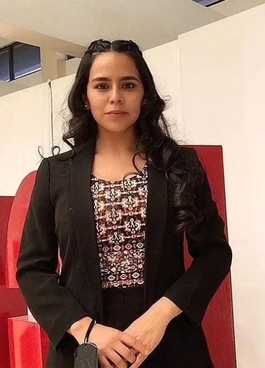 A woman with long, dark hair is standing indoors. She is wearing a black blazer over a patterned top and holding a small object in her hands. The background features white walls and a large red letter "E".