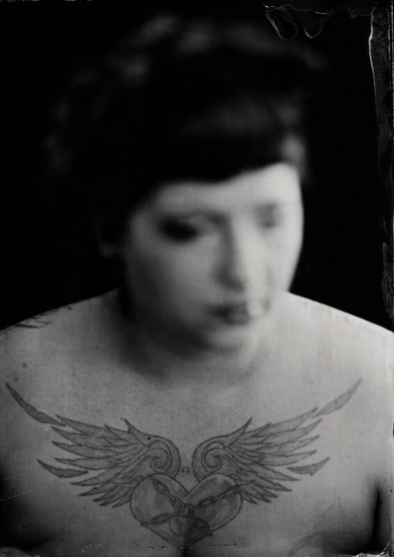 Black and white photo of a person with short, dark hair and blurred facial features. The focus is on their chest, revealing a prominent tattoo of a heart with wings and ornate detailing.
