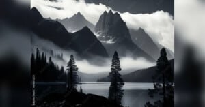 A black-and-white landscape photo depicts a misty lake surrounded by tall pine trees and dramatic mountain peaks with low-hanging clouds. The serene and atmospheric scene captures the rugged beauty of nature.
