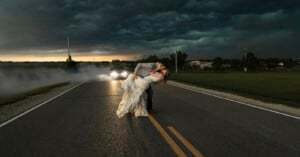 A couple shares a romantic kiss in the middle of a deserted road under a dark, stormy sky. The groom dips the bride as they embrace, while car headlights illuminate them from behind, creating a dramatic and cinematic atmosphere.