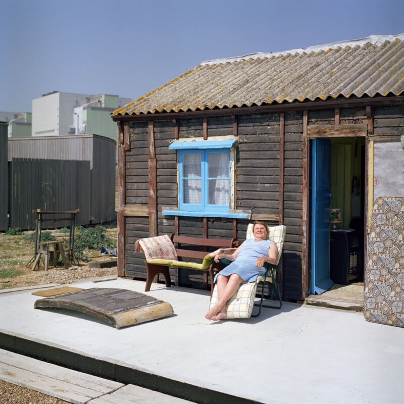 An elderly woman, wearing a light blue dress, lounges on a reclining chair outside a rustic wooden cabin. The cabin has a blue window frame and a corrugated roof. Various outdoor items are scattered nearby, and a building complex is visible in the background.
