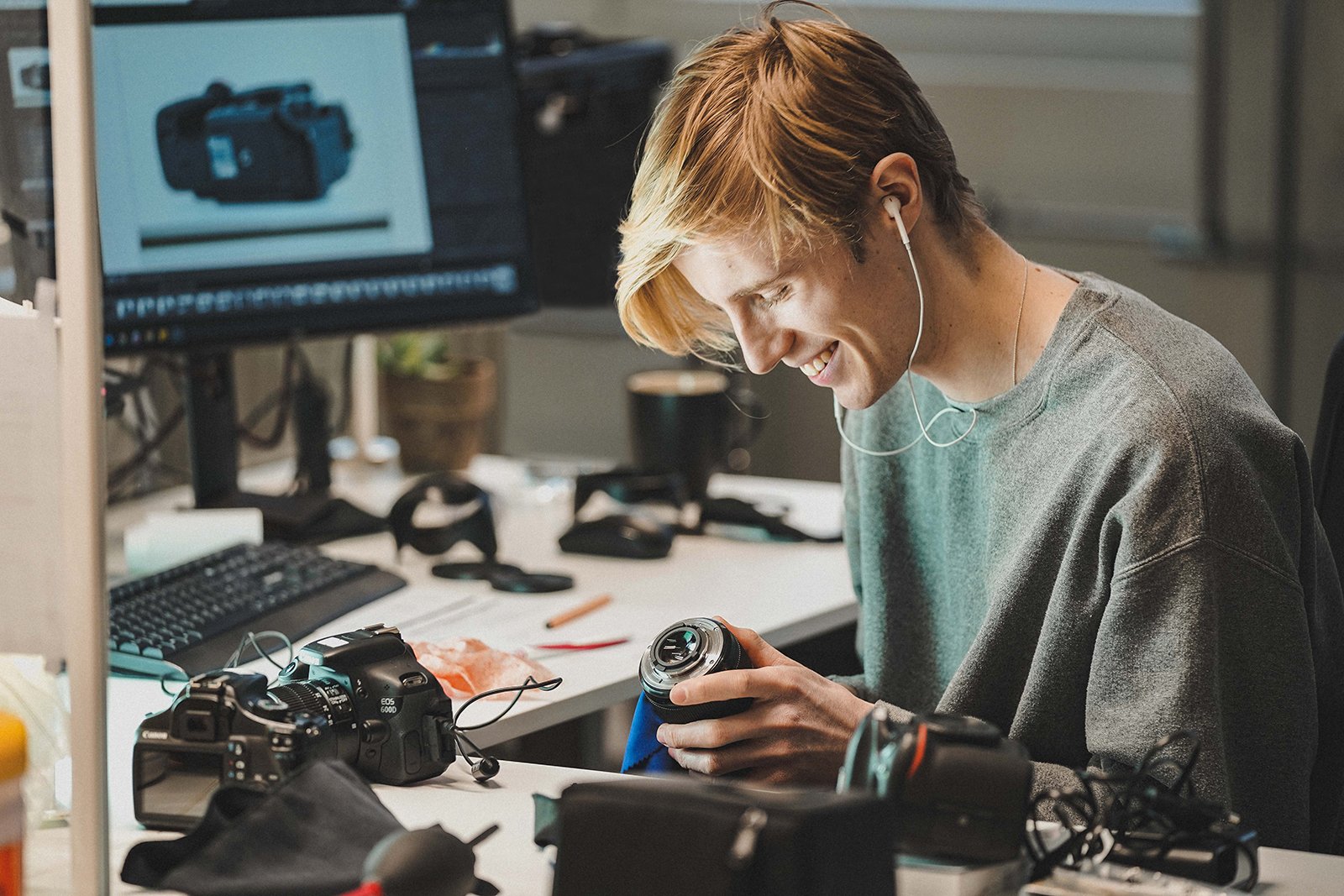 A person with earphones sits at a cluttered desk, smiling and inspecting a camera lens. A computer monitor displaying a camera is in the background. Various camera equipment and accessories are scattered across the desk.