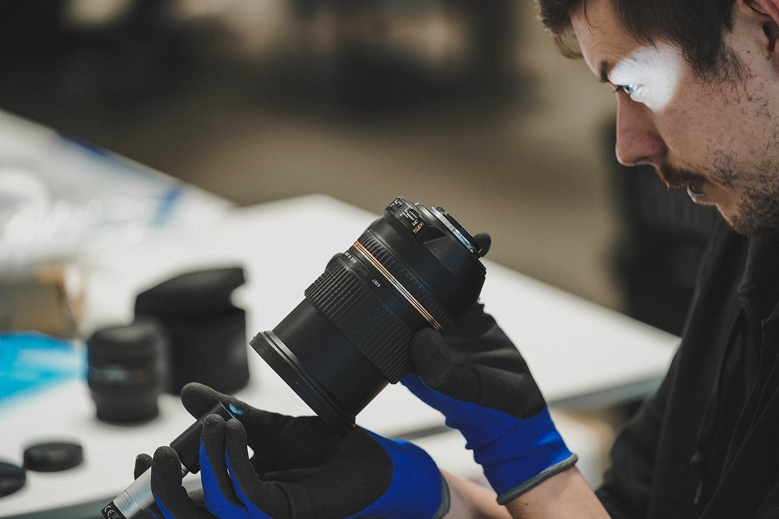 A person wearing blue and black gloves closely inspects a camera lens under a bright light. Various camera equipment is visible on the table in the background.