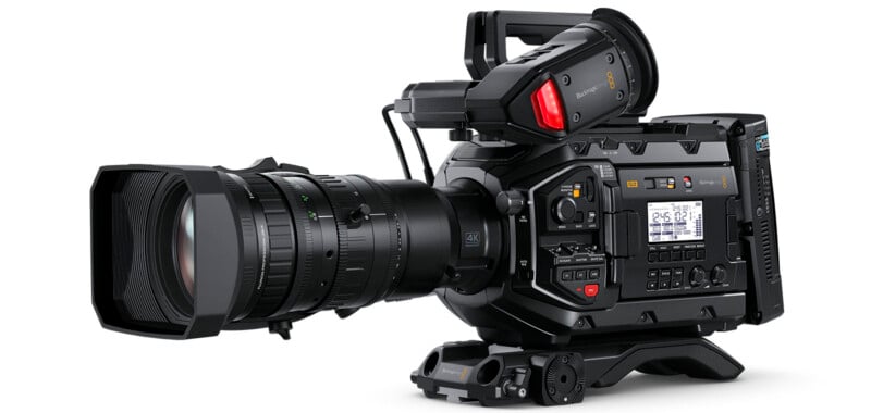 A professional 4K video camera with a large lens and numerous controls on the side. The camera features an electronic viewfinder, a top handle, and various buttons and dials for adjusting settings. It is set on a tripod base.