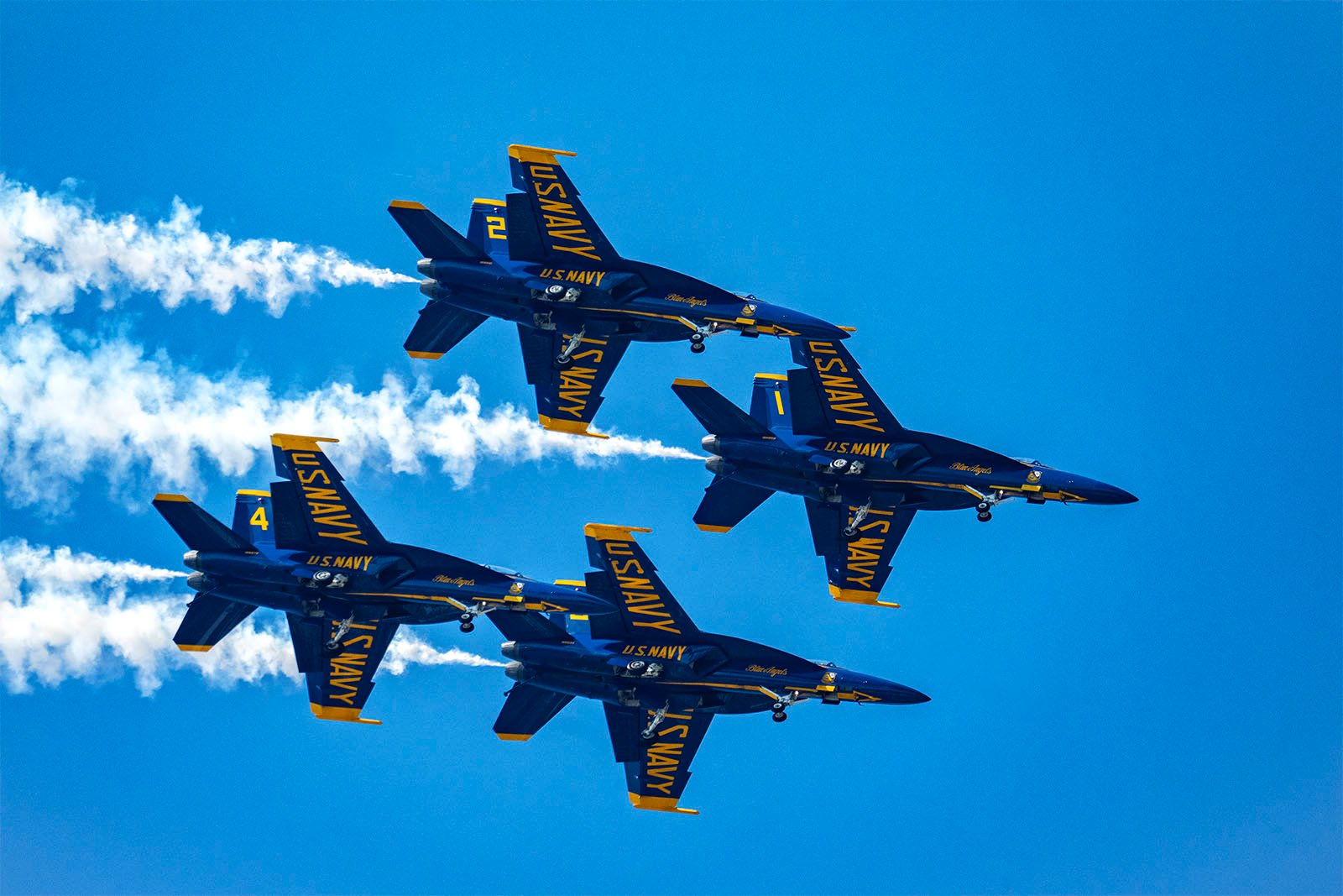 Four U.S. Navy Blue Angels jets fly in a tight formation against a clear blue sky. The planes trail white smoke and are painted navy blue with yellow accents, with "U.S. Navy" written prominently on the undersides of the wings and fuselage.