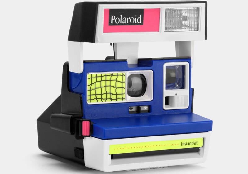A vintage Polaroid instant camera with a blue and white body and a pink Polaroid logo at the top. The camera features a yellow grid pattern on the flash area, a viewfinder, and a red button on the left side. The front has "InstantArt" written in yellow text.