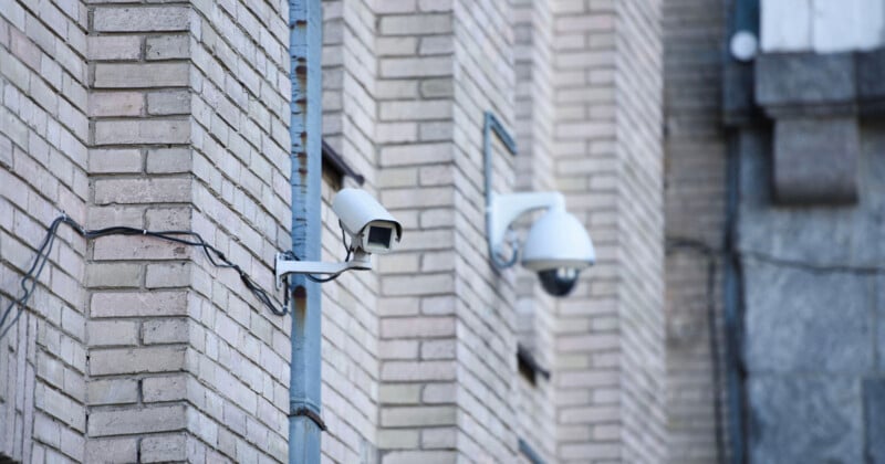Two security cameras are mounted on the corner of a brick building. One camera is in a rectangular housing, while the other is in a dome-shaped enclosure. Both cameras are connected by visible wiring.