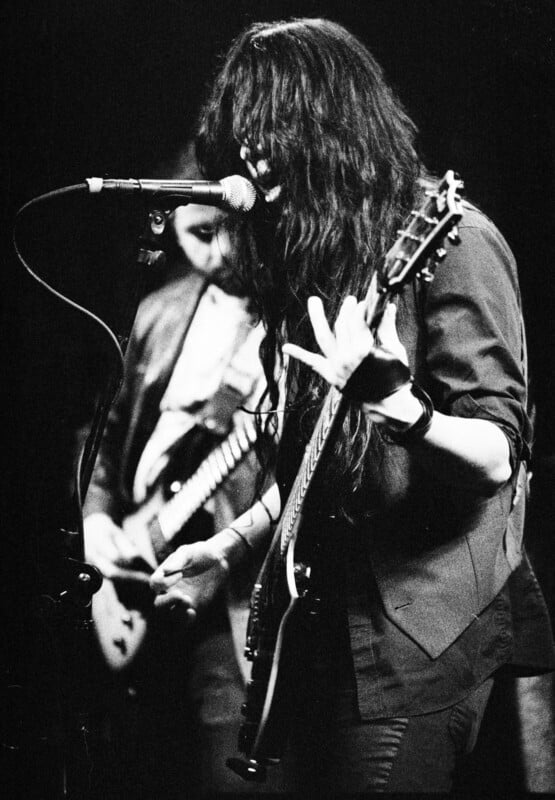 Black and white photo of a long-haired musician singing into a microphone while playing an electric guitar. Another musician is playing guitar in the background. The scene is dimly lit, giving it an intense, moody atmosphere.