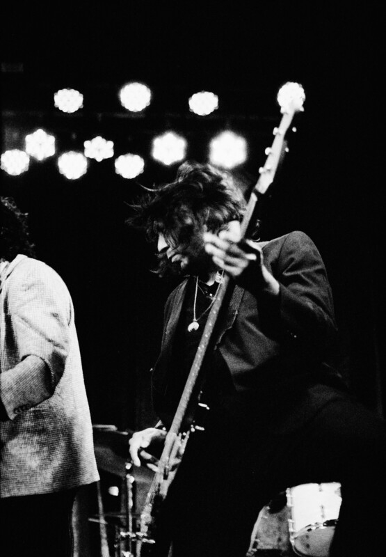 A black-and-white photo of a bass guitarist playing on stage under dramatic lighting. They are dressed in dark attire and have long hair. Another band member, only partially visible, is to their left. In the background, stage lights and a drum set can be seen.