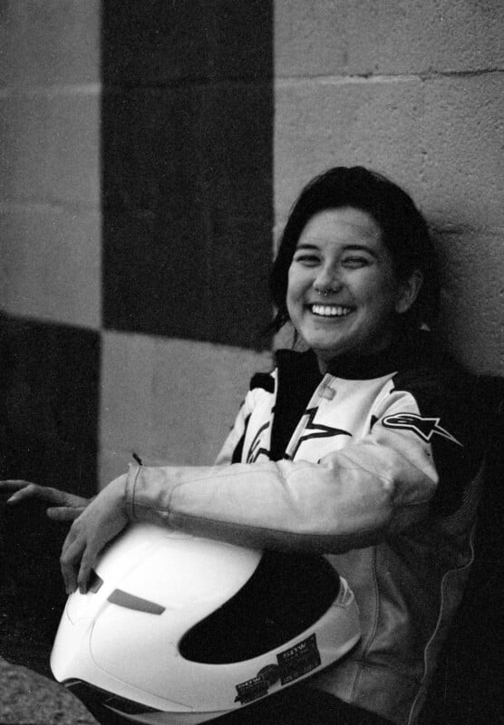 A black and white photo of a smiling person sitting against a patterned wall. The person is wearing a motorcycle jacket and holding a helmet. They appear relaxed and happy.