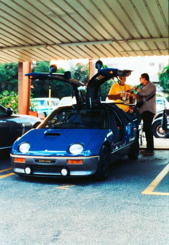 A blue car with gull-wing doors open is parked under a carport. Two individuals stand nearby, engaged in conversation. The background includes other vehicles and greenery. The car's distinctive headlights and the unique door design are prominently visible.