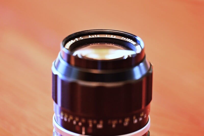 Close-up image of a vintage camera lens with text "Auto 1:2.8 f=135mm" visible on the rim. The lens has a metallic, slightly worn finish and is placed against a blurred, warm-toned background, creating a shallow depth of field effect.