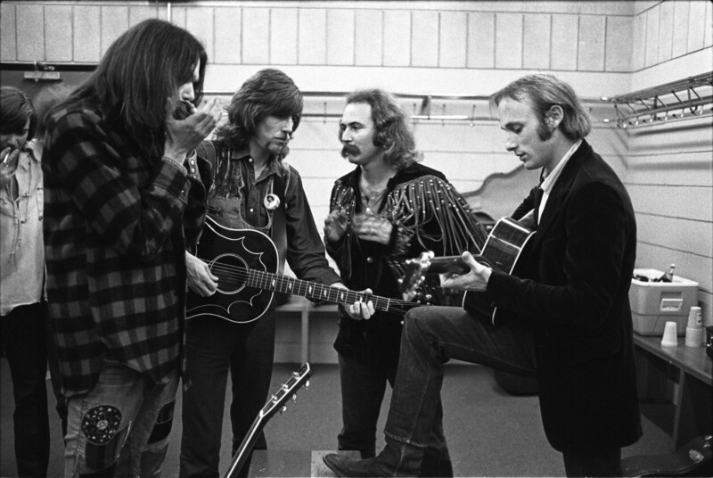 Four men are standing in a room, two playing guitars and two looking on. One of the guitar players has a foot raised on a bench. The room appears to be a dressing or rehearsal area, with visible shelves and a table with bottles and cups.