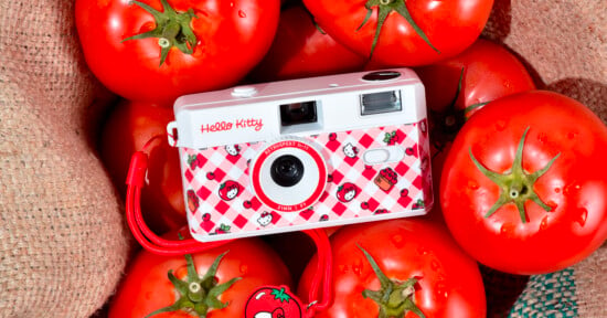 A Hello Kitty-themed camera is placed on a pile of fresh red tomatoes. The camera has a white and red checkered design with multiple Hello Kitty faces and a red wrist strap. A tomato-themed charm is attached to the strap.