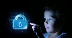 A child touches a digital padlock icon on a dark screen with a circuit board background, symbolizing cybersecurity. The lock icon emits a blue glow, illuminating the child's face.