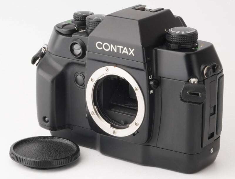 A black Contax SLR camera body with no lens attached is placed on a white background. The lens mount is exposed, and the lens cap is lying next to the camera. The camera features various dials and buttons on its top and sides.
