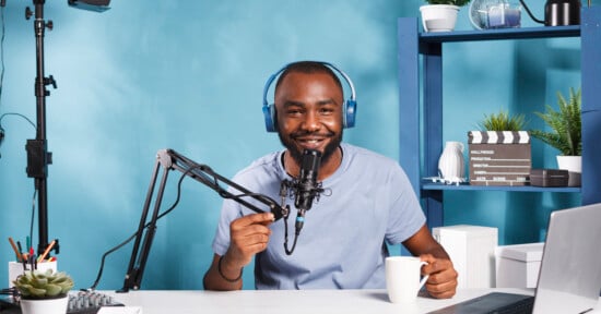 A man is sitting at a desk with a microphone and headphones, holding a white mug and smiling. The background is blue with a shelving unit holding plants, a clapperboard, and decorative items. Various electronic equipment is on the desk.