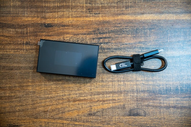 A black rectangular portable hard drive, wrapped in a transparent plastic cover, is placed on a wooden surface. Next to it is a black USB-C cable secured with a velcro strap.