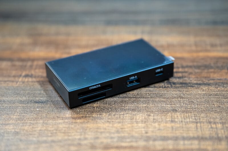 A small, rectangular USB hub with several ports, including a USB-A, a USB-C, a CF/CFast card slot, and an SD card slot, sits on a wooden surface. The device is black with labels indicating the different ports.