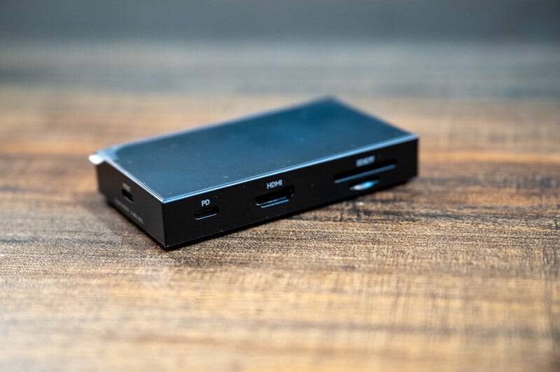 A black multi-port adapter with several connections, including a USB-C port labeled "PD", an HDMI port, and an SD card slot, sits on a wooden surface. The adapter is compact and rectangular in shape.