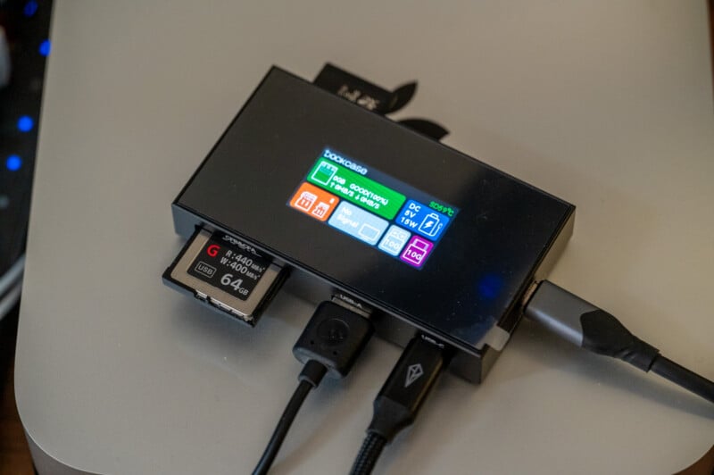 A black device with multiple ports is shown. It has a small color LCD screen displaying various data icons and text. Several cables, including a USB cable, are connected to it, along with a 64GB SanDisk SD card inserted into one of the slots.