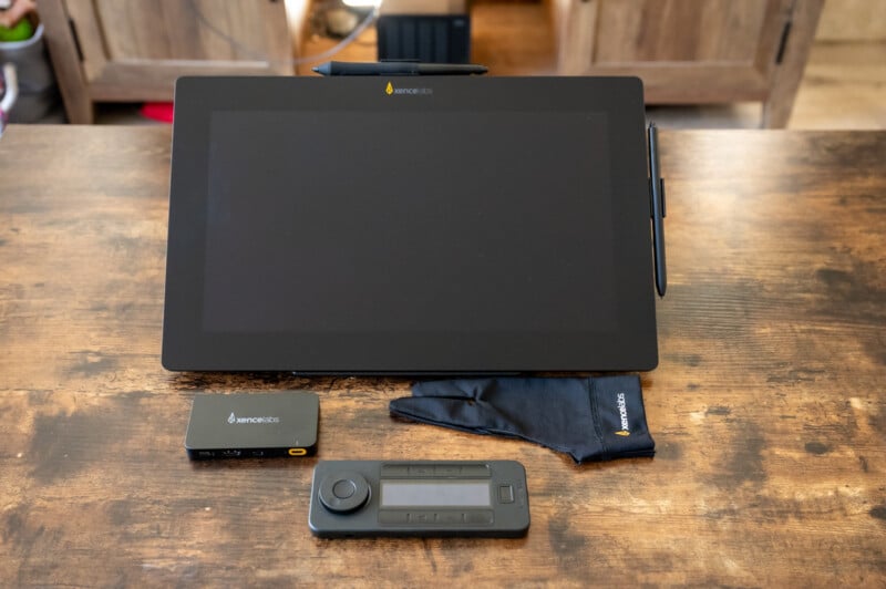 A digital drawing tablet with a stylus attached to the side is placed on a wooden table. Accompanying the tablet are a black glove, a rectangular accessory, and a small wireless remote with buttons and a wheel, all branded with the name "Xencelabs.