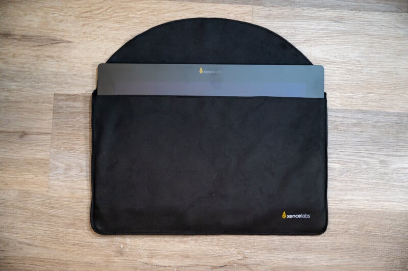 A sleek, black laptop with a visible logo is partially inserted into a matching black sleeve case. The sleeve and the laptop both have identical logos featuring a small, stylized flame icon. The items are placed on a light wooden surface.