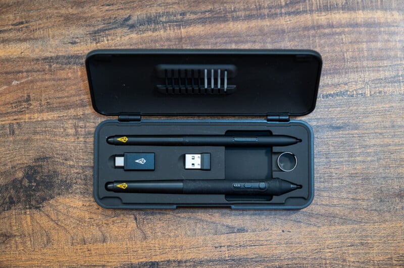 A black plastic case with a set of digital art tools is open on a wooden surface. Inside the case are a digital pen, a pen holder, a USB adapter, a ring-like tool, and extra pen nibs. The case is neatly organized with foam cutouts for each item.