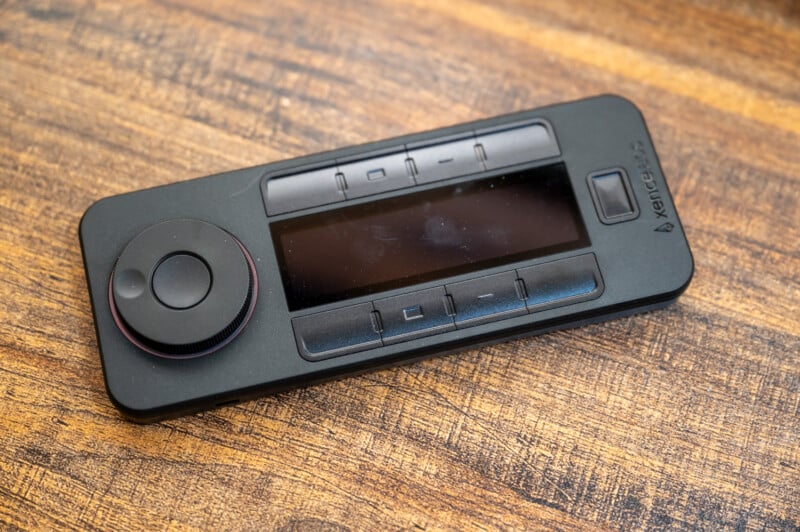 A black multimedia remote control with a screen in the center and buttons arranged around it is placed on a wooden surface. The remote has circular dials and navigation buttons, with a brand name visible on the top right corner.