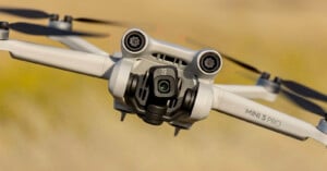 A close-up shot of the DJI Mini 3 Pro drone in flight, showing its front-facing camera, gimbal, and propellers. The background is a blurred, light brown landscape, highlighting the drone's details. The model name "MINI 3 PRO" is visible on one of its arms.