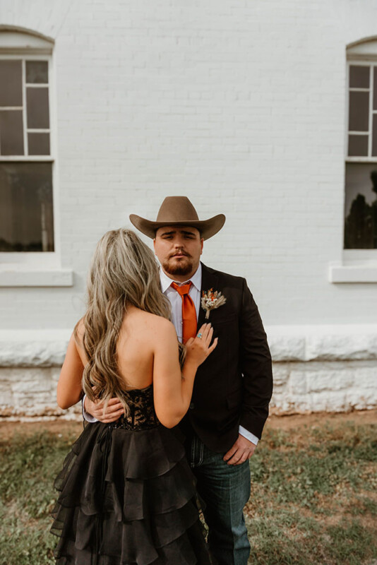 A man wearing a cowboy hat, a dark suit, orange tie, and a boutonnière stands outside against a white brick wall, with a woman's back facing the camera. The woman, with long blonde hair, wears a black strapless dress with a ruffled skirt.