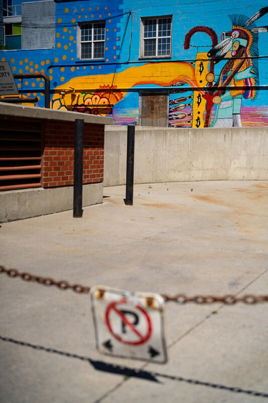 A vibrant mural featuring an indigenous character in traditional attire with a headdress, colorful patterns, and symbols is painted on the side of a building. The foreground shows a concrete barrier and a chain with a "No Parking" sign.