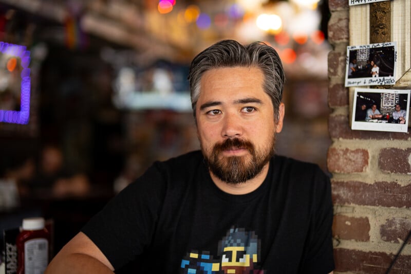 A man with short, graying hair and a beard sits casually beside a brick wall adorned with polaroid photos. He is wearing a black T-shirt with pixel art graphics on it. The background is softly lit with colorful lights, creating a cozy atmosphere.