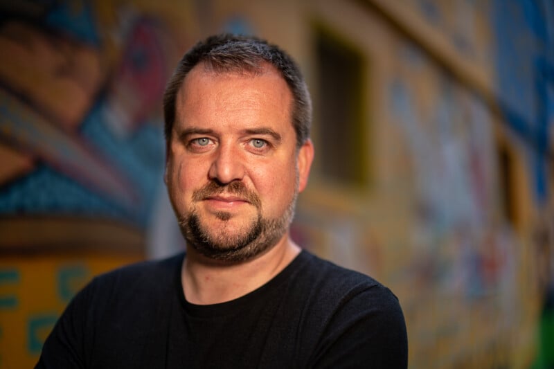 A bearded man with short hair wearing a black t-shirt stands with his arms crossed in front of a colorful graffiti wall. He looks directly at the camera with a neutral expression. The graffiti features abstract shapes and vibrant colors.
