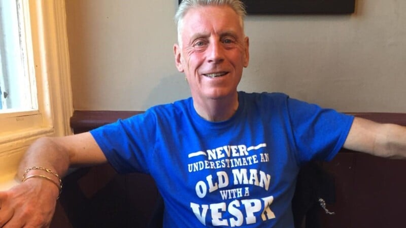 A smiling man with short, gray hair is sitting by a window. He is wearing a bright blue T-shirt that says, "Never underestimate an old man with a Vespa" in white text. He has one arm resting on the back of the bench and the other arm on the table.