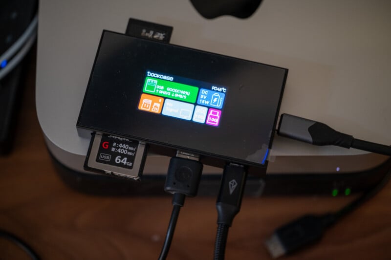 A close-up of a tech device displaying performance metrics on a small screen. Several ports are visible, with various cables and a 64GB SD card connected. The device is placed on a desk with a computer partially visible in the background.