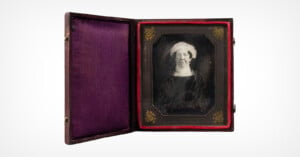 A framed, black and white photograph of an elderly woman wearing a headscarf, displayed in an antique case with a purple velvet lining and red trim. The case is open, showcasing the portrait.