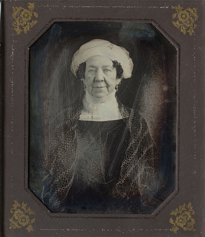 A sepia-toned portrait of an older woman with dark curly hair, wearing a white bonnet and a dark dress with a lace shawl. The image is framed in a decorated, antique-looking border with gold floral designs in the corners.