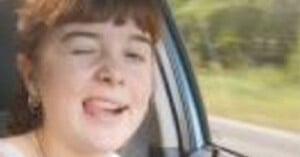 A person with light skin and brown hair is sitting in the driver's seat of a car, sticking their tongue out and winking. The car window is partially visible, and a blurred green landscape can be seen outside.
